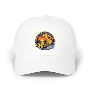 May include: White baseball cap featuring a colorful graphic of a leaping fish with the text "REEL LIFE" in a stylized font. The graphic includes a sunset scene with water and a fishing hook. The cap has a curved brim.
