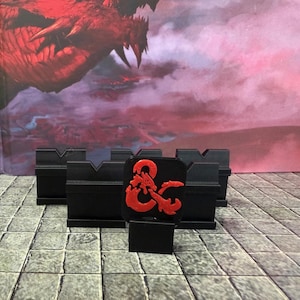May include: Black tabletop game terrain pieces with a red and black logo. The pieces are designed to hold game tiles and miniatures. The background features a red dragon illustration.