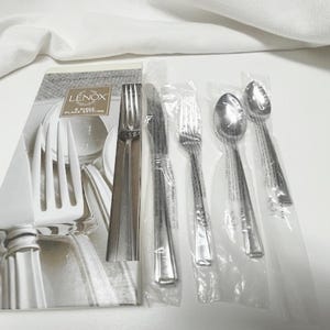 May include: A Lenox 5-piece place setting of stainless steel silverware, including a fork, knife, spoon, and teaspoon, is displayed in clear plastic packaging. The set is accompanied by a Lenox brochure.