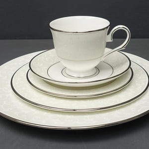 May include: A white teacup and saucer set with a silver trim, resting on a stack of matching plates. The plates have a delicate, swirling pattern. The set includes a dinner plate, salad plate, and a smaller saucer.