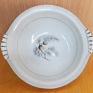 May include: White serving bowl with a pine cone and branch design. The bowl has a silver rim and handles on either side. The pine cone is brown and the branches are gray and blue. A decorative piece for serving food.