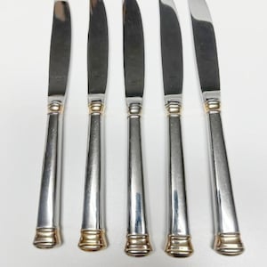 May include: Five stainless steel butter knives with silver blades and handles, accented with gold-colored bands at the base and near the blade. The knives are arranged vertically against a white background, showcasing their elegant design.