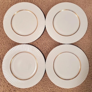 May include: Four white ceramic plates with a ridged edge design. Each plate has a gold border with a repeating geometric pattern. The plates are arranged in a square formation, suitable for a formal dining setting.