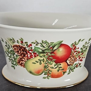 May include: White ceramic bowl with a flared shape and a gold rim. The bowl is decorated with a colorful illustration of apples, pears, and pinecones, surrounded by green leaves and red berries. A decorative piece.