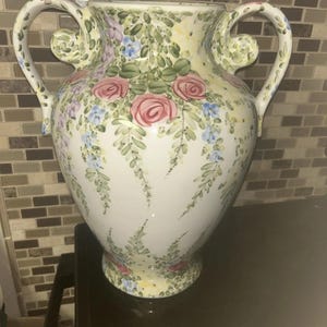 May include: A white ceramic vase with two curved handles, featuring a hand-painted floral pattern of pink roses, blue flowers, and green foliage. The design covers the exterior surface, showcasing a symmetrical arrangement of blossoms and leaves.