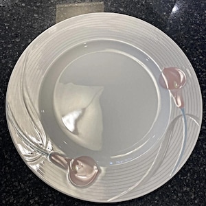 May include: A white ceramic dinner plate with a floral design. The plate features a raised, stylized calla lily design in pink and gray. The plate has a ridged border and is suitable for serving meals.