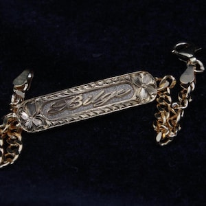 May include: A gold-toned metal identification bracelet featuring a central rectangular plate with the name Bilge engraved in script. The plate is bordered by a textured pattern and floral motifs at each end, connected to a linked chain with a clasp.