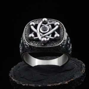 May include: A silver-toned ring featuring a prominent atomic symbol design on its face. The center of the atom is set with a small, dark, circular stone. The band and the square-shaped face are decorated with intricate, dark, engraved scrollwork.