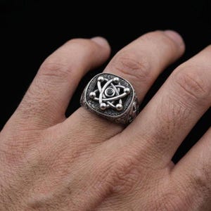 May include: A silver ring featuring a central atomic symbol design with a small black stone at its core. The ring has an oxidized finish with intricate engraved patterns along the band and the square-shaped face surrounding the atom motif.