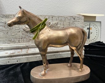 May include: A polished brass horse figurine standing on an oval base. The horse features a smooth, metallic finish and is adorned with a small, bright green velvet ribbon tied around its neck. The sculpture is captured in a side profile view.