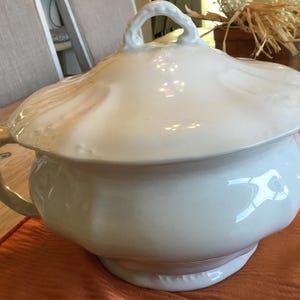 May include: A white ceramic chamber pot with a lid. The pot has a handle and a decorative rim. The lid has a loop handle. The pot is sitting on an orange surface.