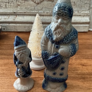 May include: Two ceramic figurines of bearded figures wearing pointed hats and long coats, one larger than the other, featuring a blue and beige glazed finish. A small, cream-colored bottle brush tree stands between the two figures.