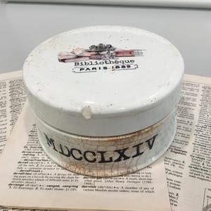 May include: A round white ceramic container with a lid featuring a graphic of books and the text "Bibliothèque PARIS 1889". The base of the container has a crackled finish and displays the black Roman numeral text "MDCCLXIV" around its side.