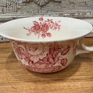 May include: A cream-colored ceramic teacup featuring a detailed red floral pattern. The exterior displays a large bouquet of flowers in a basket, while the interior rim is decorated with a smaller floral sprig. It has a single curved handle.