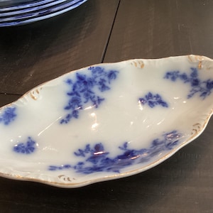 May include: An oval porcelain dish featuring a white base with blue floral patterns and gold-painted scalloped edges. The dish has a shallow, elongated shape with a smooth, glossy finish and delicate blue botanical motifs scattered across the surface.