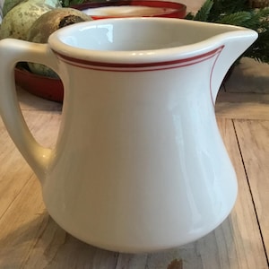 May include: A white ceramic pitcher with a curved handle and a red stripe around the rim. The pitcher has a rounded body and a spout for pouring. It is a classic kitchenware item.