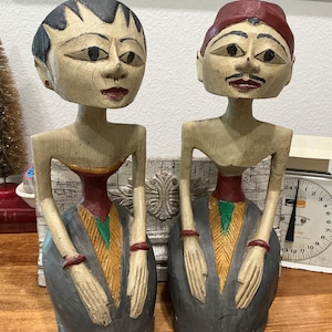 May include: Two hand-carved wooden figurines featuring stylized human figures with elongated torsos. The left figure has black hair, while the right figure wears a red headpiece. Both have painted facial features, red bracelets, and garments in shades of gray, red, green, and yellow with a distressed, crackled finish.