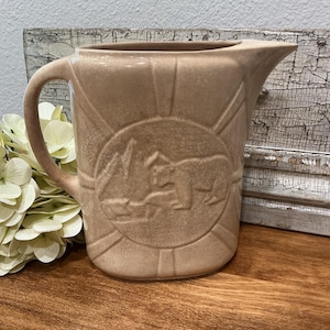 May include: A tan ceramic pitcher featuring a raised circular relief design on the front. The relief depicts a bear standing near a mountain landscape. The pitcher has a smooth handle, a wide spout, and a subtle crackled glaze finish throughout.