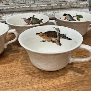 May include: A set of three white ceramic teacups with embossed leaf patterns on the exterior. Each cup features a colorful illustration of a different bird perched on a branch printed on the interior base of the bowl.
