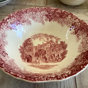 May include: A round, antique serving bowl with a cream-colored interior and a burgundy floral and landscape design. The rim has a scalloped edge with a floral pattern. The center features a detailed illustration of a building and figures.