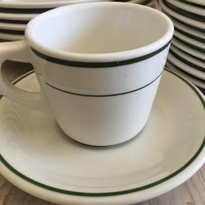 May include: White ceramic teacup and saucer set with a dark green trim. The teacup has a handle and sits on a matching saucer. Several other cups and saucers are stacked in the background.