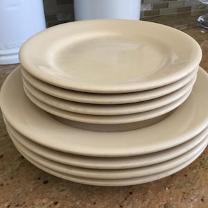 May include: A stack of off-white ceramic plates. The plates are round with a slightly raised edge. There are seven plates in total, stacked in two groups. The plates are on a speckled countertop. The plates are a neutral color.