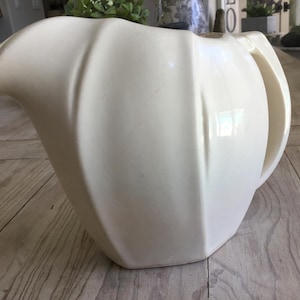 May include: Cream-colored ceramic pitcher with a curved spout and a large, arched handle. The pitcher has a ribbed design on the body and a geometric base. This vintage-style pitcher is suitable for serving beverages.