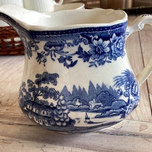 May include: White and blue ceramic pitcher with a detailed landscape and floral design. The pitcher features a curved handle and spout, with a blue and white color scheme. The design includes trees, mountains, and buildings.