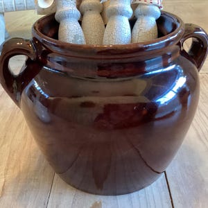 May include: A glossy, dark brown ceramic pot with two handles. The pot contains several white and beige wooden utensils. The pot sits on a light-colored wooden surface. The pot has a wide opening and a rounded body.