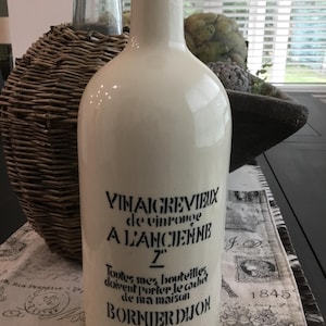 May include: A cream-colored ceramic bottle with a narrow neck and a rounded body. The front features black printed text that reads VINAIGRE VIEUX de vin rouge A L'ANCIENNE, followed by additional French text and the brand name BORNIER DIJON.