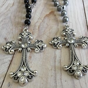 May include: Two decorative silver crosses with clear crystal accents and black beaded chains. The crosses have ornate designs and are displayed on a light wood-grain surface. The jewelry is suitable for religious occasions.