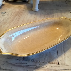 May include: A long, oval-shaped serving dish with a cream-colored interior and a gold-colored trim. The dish has a decorative, scalloped edge and is set on a light-colored wooden surface.