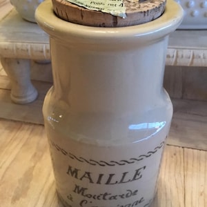 May include: A beige ceramic jar with a cork lid. The jar has the word "MAILLE" and "Moutarde à l'ancienne" printed on it. The cork lid has a label with text. The jar is approximately 6 inches tall.
