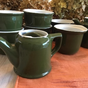 May include: A collection of green ceramic kitchenware with white trim. The set includes a small watering can, bowls, and mugs. The items are arranged on a wooden surface with a dried floral arrangement in the background.
