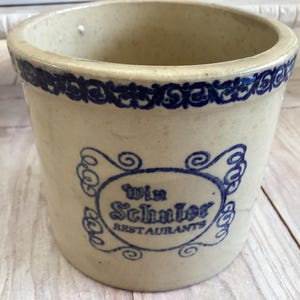 May include: A vintage, off-white stoneware crock with a blue decorative band around the rim. The crock features a blue oval emblem with the text "Win Schuler Restaurants". The crock is cylindrical and sits on a light-colored wooden surface.