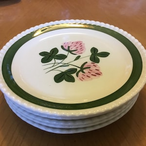 May include: A stack of white ceramic plates with a green border and a gold rim. Each plate features a pink and green floral design in the center. The plates have a textured, beaded edge.