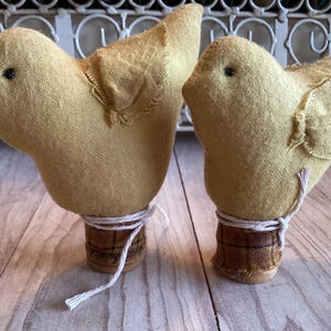 May include: Two handmade yellow fabric bird figurines. Each bird has a small black eye and is attached to a cork base with twine. The birds are a charming decorative item.