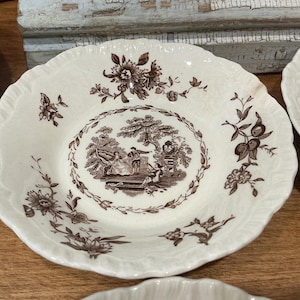 May include: A cream-colored ceramic bowl with a scalloped edge, featuring a brown transferware pattern. The center depicts a pastoral scene with figures, surrounded by a floral wreath, while the rim is decorated with brown floral motifs.
