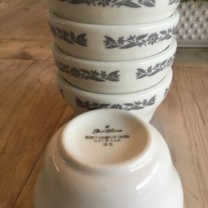 May include: A stack of white ceramic bowls with a gray floral design around the rim. The bowls are stacked on top of each other, with one bowl turned over, revealing the maker's mark on the bottom.