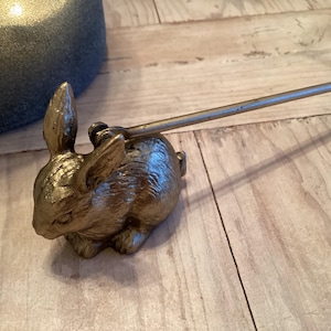 May include: A gold-colored bunny-shaped bottle stopper with a long, thin handle. The bunny is detailed with visible fur texture and is positioned on a light-colored wooden surface. The stopper is designed to fit into a bottle.
