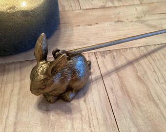 May include: A gold-colored bunny-shaped bottle stopper with a long, thin handle. The bunny is detailed with visible fur texture and is positioned on a light-colored wooden surface. The stopper is designed to fit into a bottle.