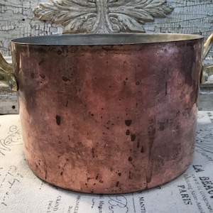 May include: A large, round copper pot with a weathered, mottled patina finish. The vessel features two sturdy, curved brass handles attached to opposite sides with metal rivets. The interior rim shows a metallic silver-toned lining.