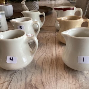 May include: A collection of cream-colored ceramic pitchers with handles and spouts, arranged on a wooden surface. Some pitchers have numbered stickers. The pitchers vary slightly in shape and size, with a few featuring a contrasting rim color.