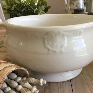 May include: Cream-colored, antique-style ceramic bowl with a handle and decorative embossed details. The bowl has a wide opening and sits on a pedestal base. A small, distressed wooden container with decorative objects is in the foreground.
