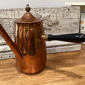 May include: A polished copper coffee pot with a conical lid, a long angled spout, and a horizontal black wooden handle. The metallic surface reflects the surroundings, and the pot features a small brass finial at the top of the lid.