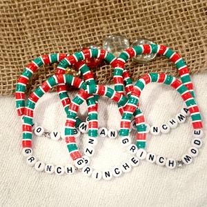 May include: Several Christmas-themed bracelets with red, green, and white disc beads. The bracelets spell out "GRINCH MODE", "GRINCH MAN", and "LOVE GRINCH". The bracelets are on a white surface with a burlap background.