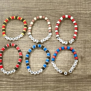 May include: Six colorful beaded bracelets with various phrases. The bracelets feature a mix of red, black, yellow, blue, and white beads. Phrases include "SINNERS", "JUKE JOINT", "BITE ME", "STACK", "SMOKE", and "BLUES BLOOD".