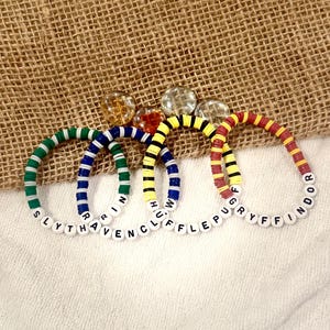 May include: Four colorful beaded bracelets with white letter beads spelling out "Slytherin", "Ravenclaw", "Hufflepuff", and "Gryffindor". The bracelets are green, blue, yellow, and red with white accents. The background is a textured tan fabric.