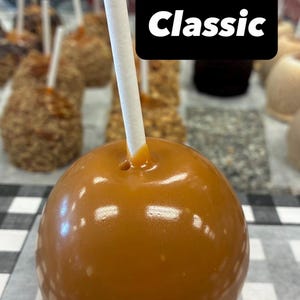 May include: A smooth, shiny caramel apple with a white stick inserted into the top center. A black rectangular sign with the white text "Classic" is positioned behind the apple, which sits among other caramel apples on a white surface.