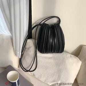 Puede incluir: Un bolso negro plisado con correas largas y finas. El bolso tiene un cierre de cordón y una forma redondeada. El bolso está sobre una superficie blanca. El fondo incluye una cortina blanca y una almohada blanca.